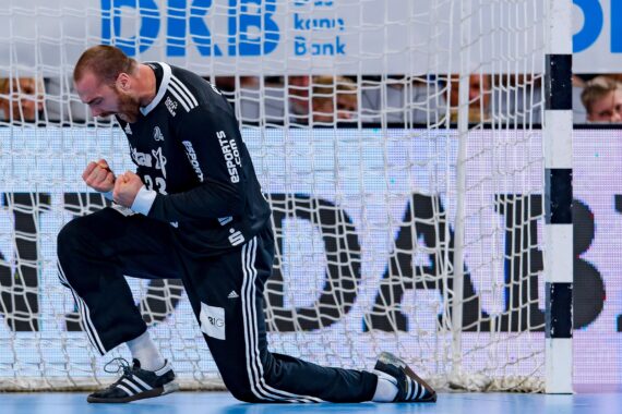 Ein männlicher Handballtorwart in einem schwarzen Trikot kniet vor dem Tor, ballt die Fäuste und schreit in Jubel aus, während im Hintergrund ein Netz und Werbebanner zu sehen sind.