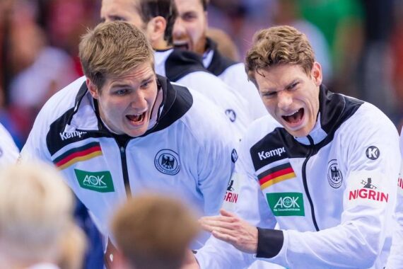 Zwei männliche Athleten in weißen deutschen Mannschaftsjacken feiern enthusiastisch, lächeln und klatschen. Im Hintergrund sind weitere Mannschaftskameraden zu sehen, die ebenfalls begeistert zu sein scheinen.