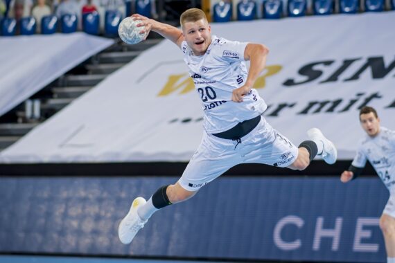 Ein männlicher Handballspieler in einem weißen Trikot springt in die Luft, um den Ball zu werfen, während eines Spiels in einer Halle mit Zuschauerplätzen im Hintergrund.