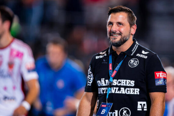 Ein lächelnder Mann in einem schwarzen Sporthemd mit Sponsorenlogos und einem Schlüsselband steht in einem Innenraum, möglicherweise bei einer Sportveranstaltung. Der Hintergrund ist unscharf, eine weitere Person in einem weiß-roten Hemd ist zu sehen.