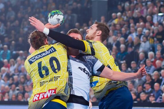 Drei Handballspieler kämpfen während eines Spiels intensiv um den Ball, wobei zwei Spieler in gelben Trikots einen Spieler in einem weiß-schwarzen Trikot blockieren. Die Szene findet in einer überfüllten Halle statt.