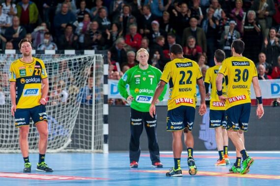 Vier männliche Handballspieler in gelben Trikots und ein Torwart in Grün stehen auf einem Hallenplatz, während im Hintergrund eine Zuschauermenge zusieht.