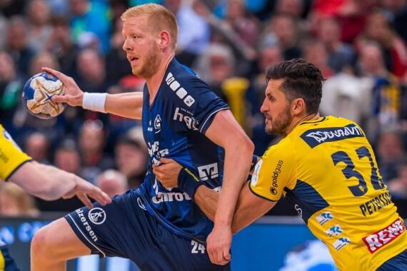 Ein Handballspieler in einem blauen Trikot versucht, sich zu befreien, während ein Verteidiger in einem gelben Trikot ihn während eines intensiven Spiels an der Hüfte festhält, mit einer unscharfen Menschenmenge im Hintergrund.