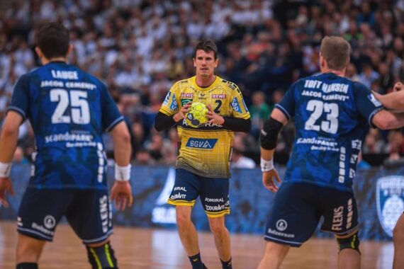 Ein Handballspieler in einem gelben Trikot hält den Ball, während sich zwei gegnerische Spieler in blauen Trikots darauf vorbereiten, ihn in einer Halle zu verteidigen, während die Zuschauer im Hintergrund zuschauen.