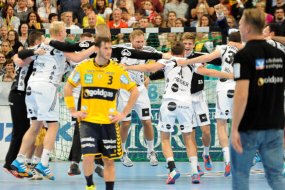 Eine Gruppe von Handballern in weißen Trikots feiert gemeinsam, während ein Spieler in einem gelben Trikot im Vordergrund enttäuscht dreinschaut. Die Zuschauer schauen von den vollbesetzten Tribünen hinter ihnen zu.