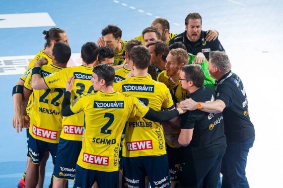 Eine Herren-Handballmannschaft in gelben und blauen Trikots versammelt sich auf einem Hallenplatz und feiert und jubelt ausgelassen. Die Trainer in schwarzen Hemden gesellen sich zu der Gruppe.