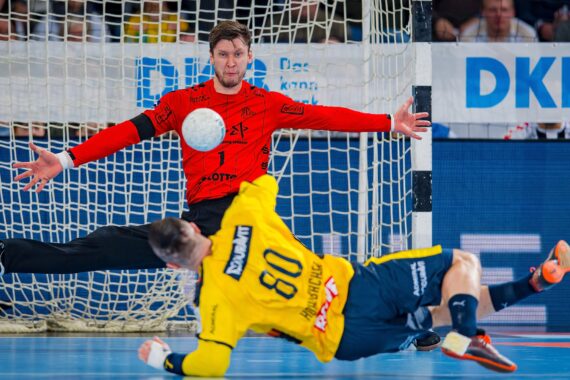 Ein Handballtorwart in einem roten Trikot streckt Arme und Beine weit aus, um einen Schuss abzublocken, während ein Spieler in einem gelben Trikot auf dem Boden rutscht und den Ball in Richtung Tor wirft. Im Hintergrund sind Zuschauer zu sehen.