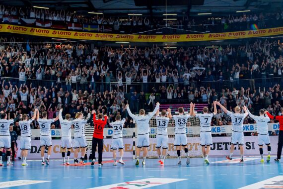 Eine Handballmannschaft in weißen Trikots steht in einer Reihe vor einer jubelnden Menge und hebt jubelnd die Arme in einer überfüllten Halle. Auch die Zuschauer heben die Arme, um ihre Begeisterung und Unterstützung zu zeigen.