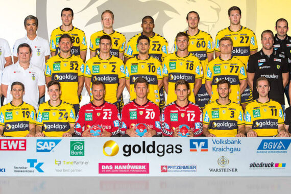 Eine Männerhandballmannschaft in gelben und schwarzen Trikots posiert mit Trainern und Betreuern vor einem Sponsorenhintergrund. Zwei Torhüter in Rot stehen in der ersten Reihe; für das Gruppenfoto stehen oder sitzen alle formell.