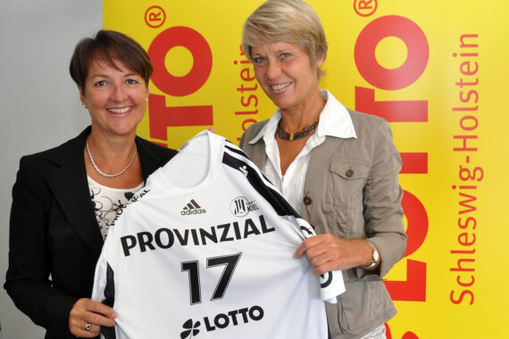 Zwei Frauen lächeln und halten ein weißes Provinzial 17-Sporttrikot vor gelben Bannern mit roten LOTTO Schleswig-Holstein-Logos.