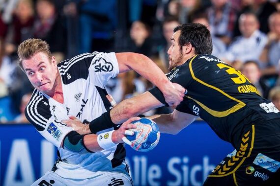 Zwei männliche Handballspieler liefern sich einen intensiven Wettkampf; der eine in einem weißen Trikot versucht, sich von einem Verteidiger in einem schwarz-gelben Trikot zu befreien, während im Hintergrund eine unscharfe Menschenmenge zu sehen ist.