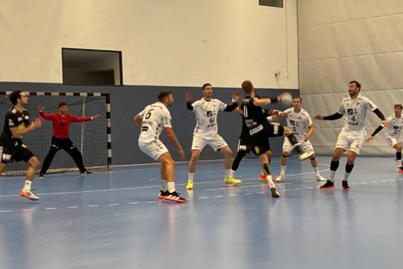 Ein Handballspieler in Schwarz springt, um den Ball in Richtung Tor zu werfen, während sechs Spieler in Weiß den Ball verteidigen und der Torwart sich darauf vorbereitet, den Schuss zu blocken.