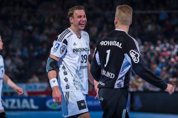Zwei männliche Handballspieler in schwarz-weißen Trikots lächeln sich auf einem Hallenplatz an, umgeben von einer unscharfen Menschenmenge im Hintergrund. Der Spieler auf der linken Seite trägt die Nummer 39, der auf der rechten Seite die Nummer 1.