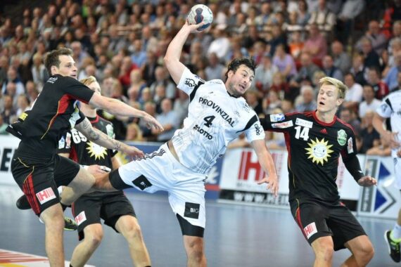 Ein Handballspieler in einem weißen Trikot springt, um den Ball zu schießen, während zwei gegnerische Spieler in schwarzen Trikots versuchen, ihn während eines Spiels zu blockieren, mit einer verschwommenen Menschenmenge im Hintergrund.
