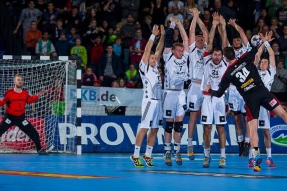 Ein Handballspieler in Schwarz springt auf das Tor zu, während fünf Verteidiger in weißen Uniformen ihn abwehren, während ein Torwart in Rot aufmerksam zusieht. Eine Menschenmenge verfolgt das intensive Spiel von der Tribüne aus.
