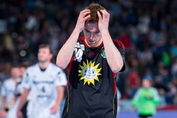 Ein Handballspieler in einem schwarzen Trikot mit einem großen Sonnenlogo steht mit den Händen auf dem Kopf und blickt offensichtlich frustriert nach unten, während im Hintergrund andere Spieler und eine verschwommene Menschenmenge zu sehen sind.