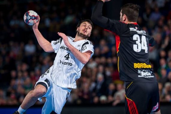 Ein Handballspieler im weißen Trikot mit der Nummer 4 bereitet sich auf den Wurf vor, während er einem Gegner im schwarzen Trikot mit der Nummer 34 während eines Spiels gegenübersteht, mit einer verschwommenen Zuschauermenge im Hintergrund.