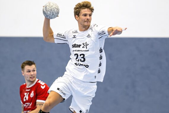 Ein männlicher Handballspieler in einem weißen Trikot springt und bereitet sich auf den Wurf vor, während ein anderer Spieler in einem roten Trikot von hinten auf einem Hallenplatz zusieht.
