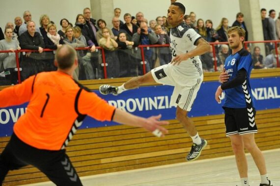 Ein Handballspieler in einem weißen Trikot springt in der Luft, um den Ball in Richtung Tor zu schießen, während ein orangefarbener Torwart sich darauf vorbereitet, den Ball zu blocken, und ein anderer Spieler in Blau in der Nähe steht. Im Hintergrund sehen Zuschauer zu.