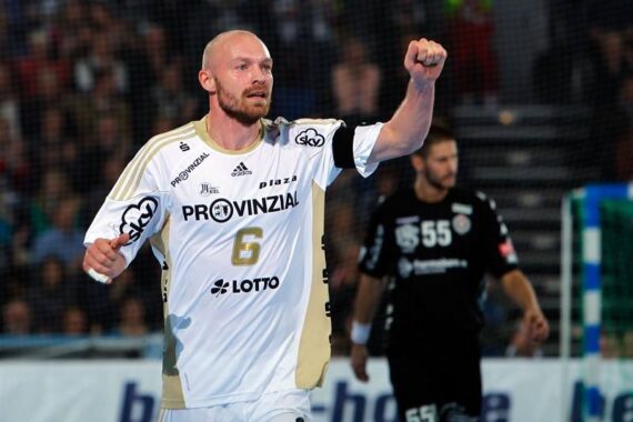 Ein männlicher Handballspieler in einem weiß-goldenen Trikot mit der Nummer 6 hebt jubelnd die Faust auf dem Spielfeld, während ein anderer Spieler in einem schwarzen Trikot im Hintergrund steht.