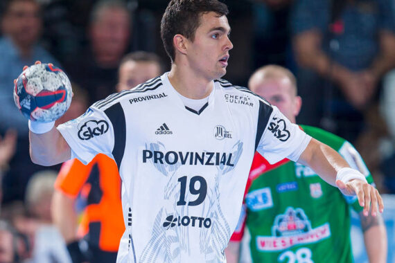 Ein männlicher Handballspieler in einem weißen Trikot mit der Nummer 19 bereitet sich darauf vor, den Ball während eines Spiels zu werfen. Im Hintergrund sind andere Spieler und ein Schiedsrichter in Orange zu sehen.
