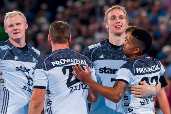 Vier männliche Handballspieler in passenden weißen und schwarzen Trikots feiern zusammen auf dem Spielfeld, lächelnd und sich umarmend nach einem Spiel, mit einer unscharfen Menschenmenge im Hintergrund.