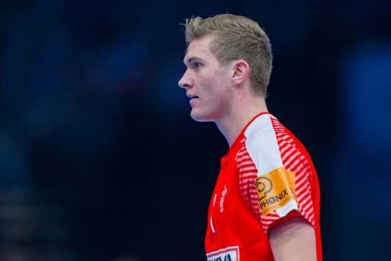 Ein männlicher Handballspieler in einem rot-weißen Trikot mit kurzen blonden Haaren steht während eines Spiels im Profil und trägt eine gelbe Kapitänsbinde. Der Hintergrund ist unscharf.