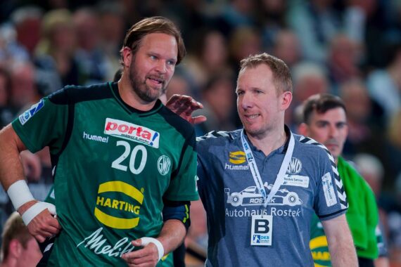 Zwei Männer stehen nebeneinander in einer Sportarena. Der eine trägt ein grünes Handballtrikot mit der Nummer 20, der andere ein graues Hemd mit einem Schlüsselband. Sie scheinen sich zu unterhalten, während im Hintergrund eine Menschenmenge zu sehen ist.