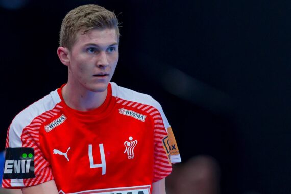 Ein männlicher Handballspieler, der ein rot-weißes Trikot mit der Nummer 4 trägt, steht konzentriert auf dem Spielfeld. Auf dem Trikot sind Logos und die Aufschrift "DENMARK" zu sehen. Der Hintergrund ist unscharf.