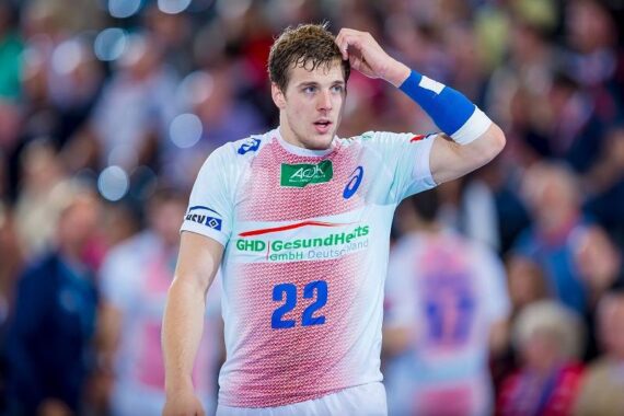 Ein männlicher Handballspieler in einem weiß-rosafarbenen Trikot mit der Nummer 22 steht auf dem Spielfeld und berührt seinen Kopf, während im Hintergrund eine unscharfe Menschenmenge zu sehen ist.