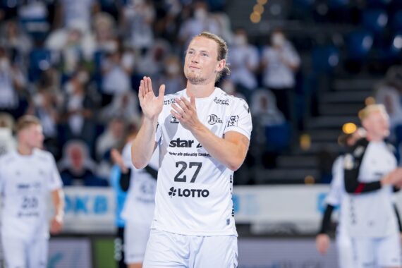 Ein Handballspieler in einem weißen Trikot mit der Nummer 27 klatscht, während er auf einem Hallenplatz steht, mit verschwommenen Mitspielern und Zuschauern im Hintergrund.