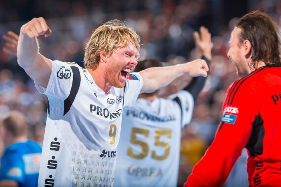 Ein männlicher Handballspieler in einem weißen Trikot feiert leidenschaftlich mit weit ausgebreiteten Armen, während er einem Mannschaftskameraden in einem roten Torwarttrikot gegenübersteht. Andere Spieler und eine jubelnde Menge sind im unscharfen Hintergrund zu sehen.