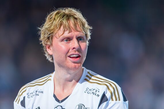 Ein blonder männlicher Sportler in einem weißen Sporttrikot mit goldenen Streifen blickt zur Seite, wirkt konzentriert und leicht errötet, der Hintergrund ist unscharf.