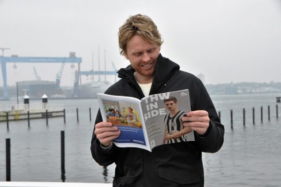 Ein Mann in einer schwarzen Jacke steht am Wasser und liest lächelnd in einer Zeitschrift. Im Hintergrund sind Boote, Docks und Kräne an einem bewölkten Tag zu sehen.