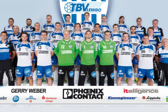 Eine Herren-Handballmannschaft in blau-weiß-grünen Trikots stellt sich für ein Gruppenfoto in drei Reihen auf, die Trainer stehen an den Seiten. Vor der Mannschaft sind die Logos der Sponsoren zu sehen.