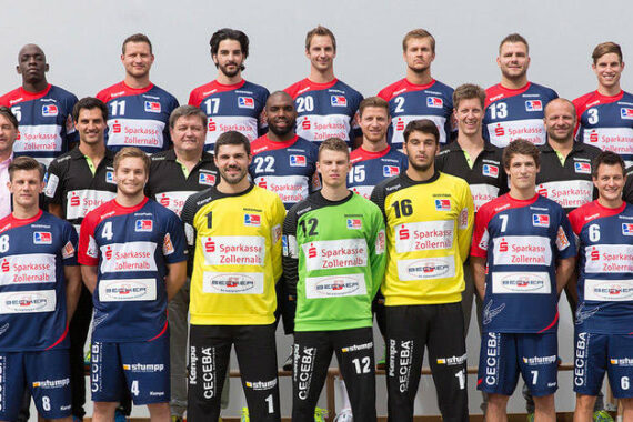 Eine Herren-Handballmannschaft posiert für ein Gruppenfoto in passenden blauen, roten und weißen Trikots. Vorne stehen zwei Spieler in gelben Torwarttrikots, hinter den Spielern Trainer und Mitarbeiter in Businesskleidung und Trainingsanzügen.