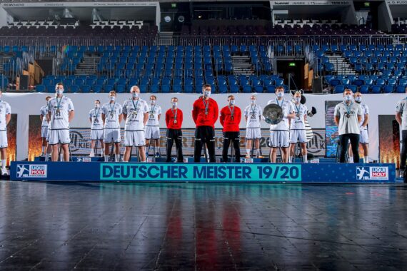 Eine Herren-Handballmannschaft steht auf einem Podium mit der Aufschrift DEUTSCHER MEISTER 19/20 in einer leeren Arena, trägt Medaillen und weiße Trikots, die Trainer tragen rote Jacken und feiern ihren Sieg.