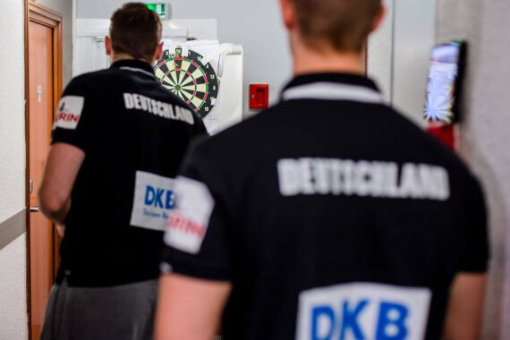 Zwei Personen in schwarzen Hemden mit der Aufschrift Deutschland stehen in einem Flur in der Nähe einer Dartscheibe, mit dem Rücken zur Kamera. Die Szene scheint bei einer Dart-Veranstaltung oder einem Dart-Wettbewerb zu spielen.