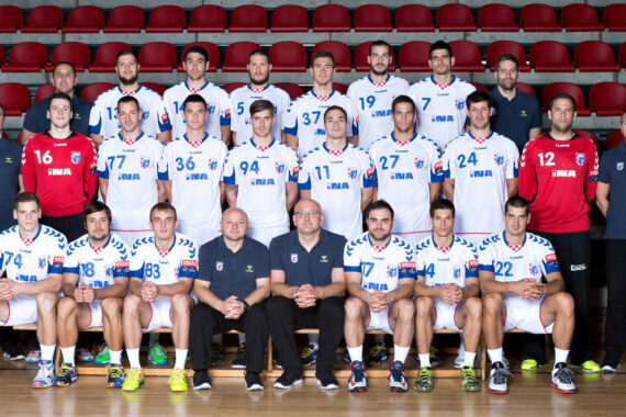 Eine Herrenhandballmannschaft stellt sich in drei Reihen in weißen Trikots mit blauen und roten Akzenten für ein Gruppenfoto auf. Trainer und Betreuer in dunkler Kleidung sitzen und stehen zwischen den Spielern. Hinter ihnen sind rote Stadionsitze zu sehen.