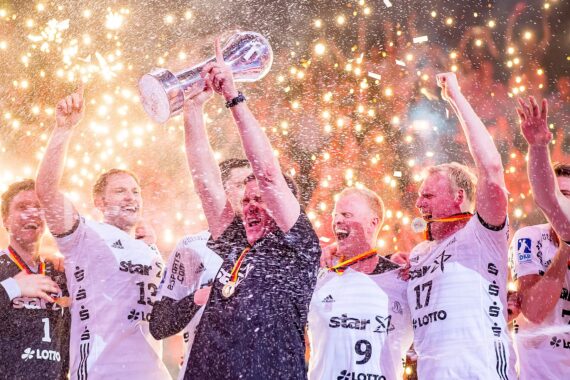 Ein jubelndes Sportteam in weißen Trikots feiert auf der Bühne, während ein Mitglied eine große Trophäe inmitten von Funken und Konfetti in die Höhe reckt, während die Mannschaftskameraden um ihn herum jubeln und lächeln.