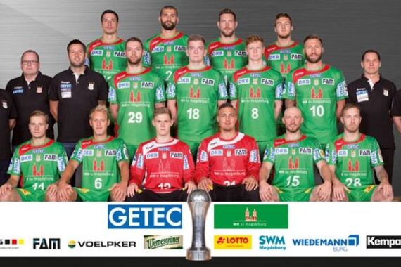 Eine Herren-Handballmannschaft posiert für ein Gruppenfoto in grünen und roten Trikots, stehend und sitzend in drei Reihen mit Trainern auf beiden Seiten. Die Logos der Sponsoren sind am unteren Rand angebracht. Vorne ist ein Pokal aufgestellt.