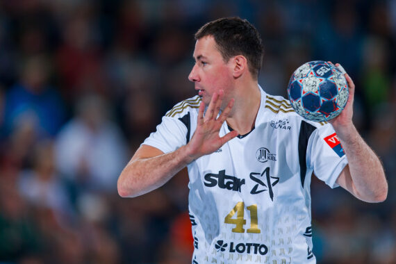 Ein männlicher Handballspieler in einem weißen Trikot mit der Nummer 41 bereitet sich darauf vor, den Ball während eines Spiels zu werfen. Er konzentriert sich mit einem erhobenen Arm und dem anderen, der den Ball hält, vor einem unscharfen Hintergrund von Zuschauern.