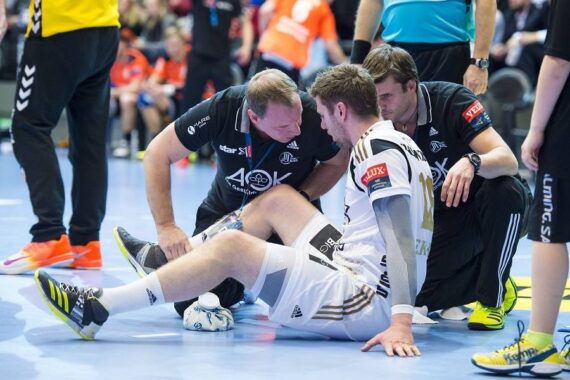 Ein männlicher Handballspieler in einem weißen Trikot sitzt auf dem Boden und scheint verletzt zu sein, während zwei Trainer in Schwarz neben ihm knien und ihm während eines Spiels Hilfe anbieten. Im Hintergrund sind unscharfe Spieler und Zuschauer zu sehen.