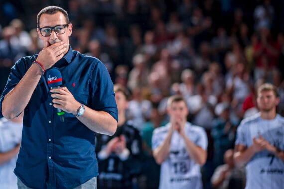 Ein Mann mit Brille und dunklem Hemd steht auf einer Bühne und hält ein Mikrofon in der Hand, während im verschwommenen Hintergrund mehrere Personen in Sporttrikots applaudieren.