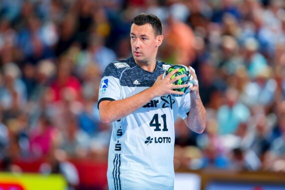 Ein männlicher Handballspieler in einem weiß-blauen Trikot, der einen grünen Handball hält und sich auf den Wurf vorbereitet, mit einer verschwommenen Menschenmenge im Hintergrund.