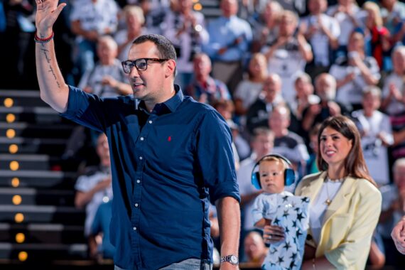 Ein Mann mit Brille und dunklem Hemd winkt einer Menschenmenge in einem Stadion zu. Neben ihm hält eine Frau in einem hellen Blazer ein Baby mit Ohrenschützern und einer Decke mit Sternenmuster. Das Publikum ist im Hintergrund unscharf zu erkennen.
