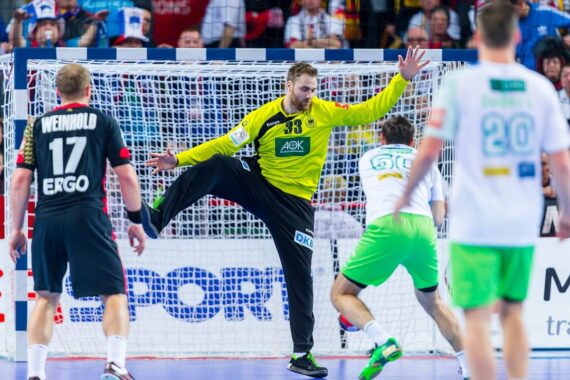 Ein Handballtorwart in einem gelben Trikot blockiert einen Schuss, während die Spieler der gegnerischen Mannschaften, die schwarze und grüne Trikots tragen, vor einer überfüllten Arena gegeneinander antreten.