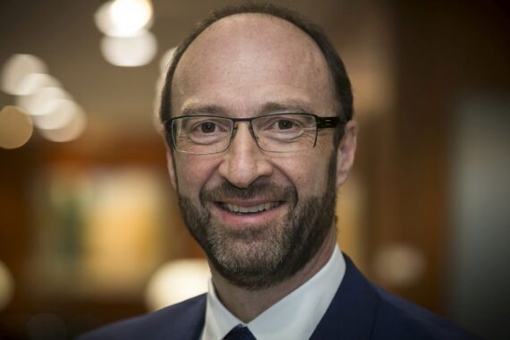 Ein Mann mittleren Alters mit kurzen braunen Haaren, einem Bart und einer Brille lächelt in die Kamera. Er trägt einen dunklen Anzug und eine Krawatte. Der Hintergrund ist sanft verschwommen und warm beleuchtet.
