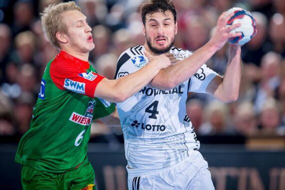 Zwei männliche Handballspieler in Aktion während eines Spiels. Der Spieler in Weiß hält den Ball, während er von einem Spieler in Grün, der einen Arm über seine Brust legt, eng verteidigt wird. Im Hintergrund ist eine unscharfe Menschenmenge zu sehen.