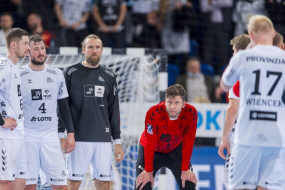 Eine Gruppe männlicher Handballspieler in weißen Trikots steht zusammen auf einem Spielfeld, wobei sich ein Spieler in einem roten Torwarttrikot nach vorne beugt. Die Spieler scheinen konzentriert zu sein, während im Hintergrund ein Netz und eine Zuschauermenge zu sehen sind.